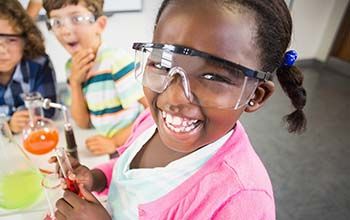 Girl smiling while holding a test tube with liquid inside. 2 other boys looking up at girl. There are 2 beakers on the desk.