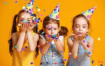 3 girls wearing birthday hats celebrating a birthday with confetti on yellow background