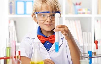 Boy holding a test tube filled with yellow liquid. He is surrounded by other beakers and test tubes with multi colored liqui